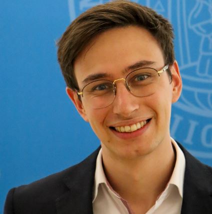 Person mit Brille, junger Mann, lächelt in Kamera, blaues Logo der Universität Wien als Hintergrund
